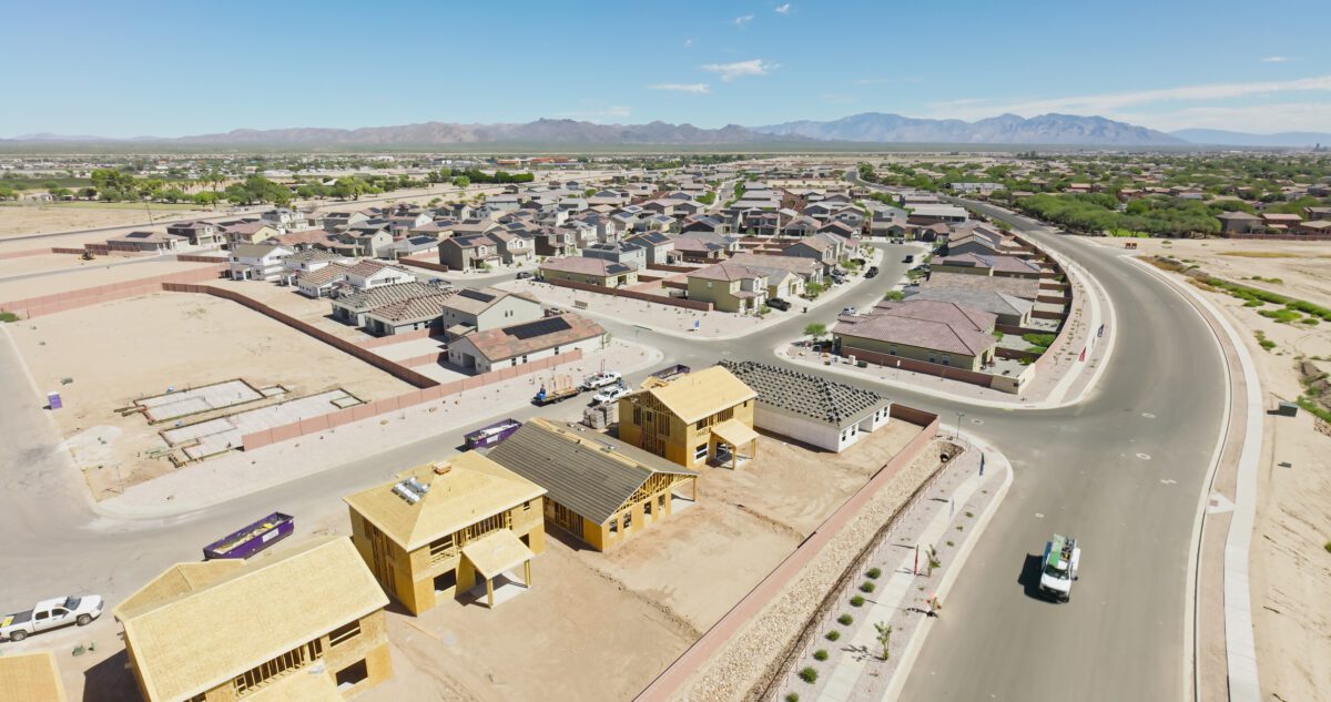 Aerial view of a residential housing development under construction in Marana, Arizona, during the day. The image shows multiple homes in various stages of completion, with wood-framed houses prominently visible in the foreground alongside finished single-family residences with tile roofs. The curved suburban streets wind through the planned community, with construction vehicles and building materials scattered throughout the active construction zone. Desert mountains are visible in the background under clear blue skies, characteristic of the Sonoran Desert landscape surrounding this growing Arizona community north of Tucson.