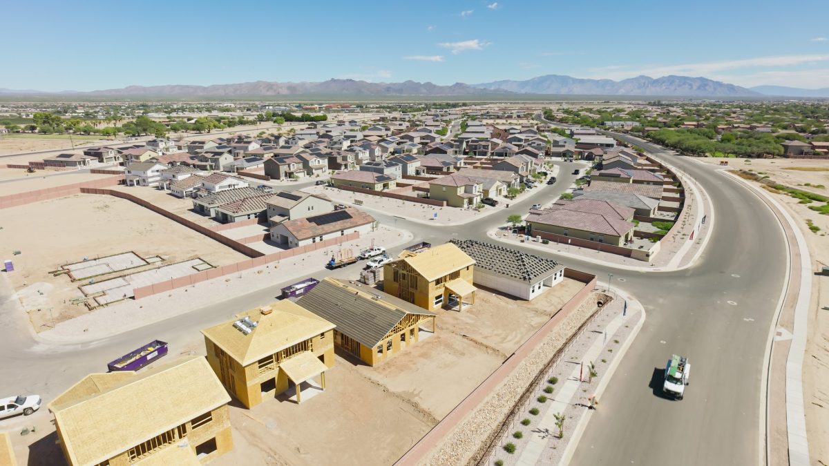Aerial view of a residential housing development under construction in Marana, Arizona, during the day. The image shows multiple homes in various stages of completion, with wood-framed houses prominently visible in the foreground alongside finished single-family residences with tile roofs. The curved suburban streets wind through the planned community, with construction vehicles and building materials scattered throughout the active construction zone. Desert mountains are visible in the background under clear blue skies, characteristic of the Sonoran Desert landscape surrounding this growing Arizona community north of Tucson.